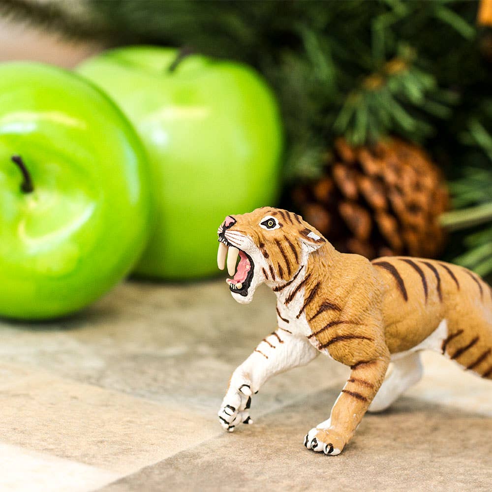 A Smilodon Saber-Toothed Cat Figurine Toy, featuring an open mouth, stands near two green apples and pinecones, with greenery in the background.