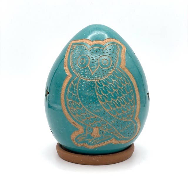 The Owl Luminary is a handcrafted, turquoise, egg-shaped ceramic with a gold-outlined owl design on its surface, set on a small round brown base.
