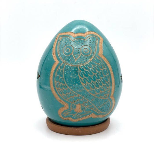 The Owl Luminary is a handcrafted, turquoise, egg-shaped ceramic with a gold-outlined owl design on its surface, set on a small round brown base.