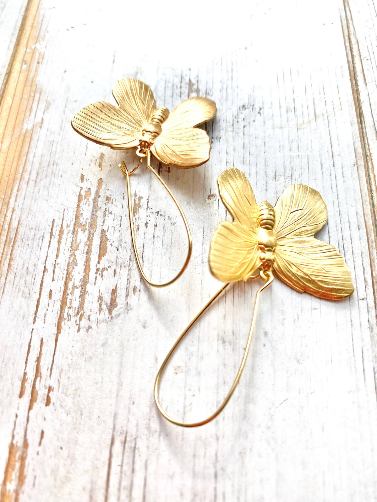 Adorn your ears with the Butterfly Earrings—two delicate, gold butterfly-shaped charms with textured wings and slim teardrop wire hooks, beautifully displayed on a whitewashed wooden surface. These stylish earrings are the perfect statement accessory for any occasion.