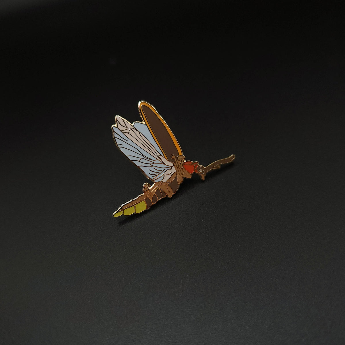 A vibrant Eastern Firefly Educational Insect Enamel Pin rests on a smooth, dark surface. This pin, inspired by the delicate form of a firefly, showcases yellow, orange, and white details with intricately designed wings—making it an ideal gift for entomology enthusiasts. Subtle glow-in-the-dark accents add a magical touch to this unique collectible accessory.