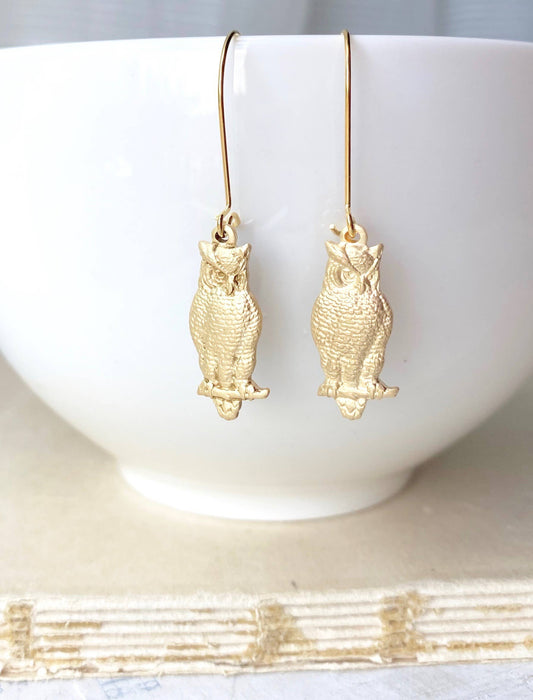 The Wise Owl Earrings, shaped like gold owls, hang in front of a white bowl and are displayed on a textured, light-colored surface. These charming earrings add a whimsical touch to any jewelry collection.