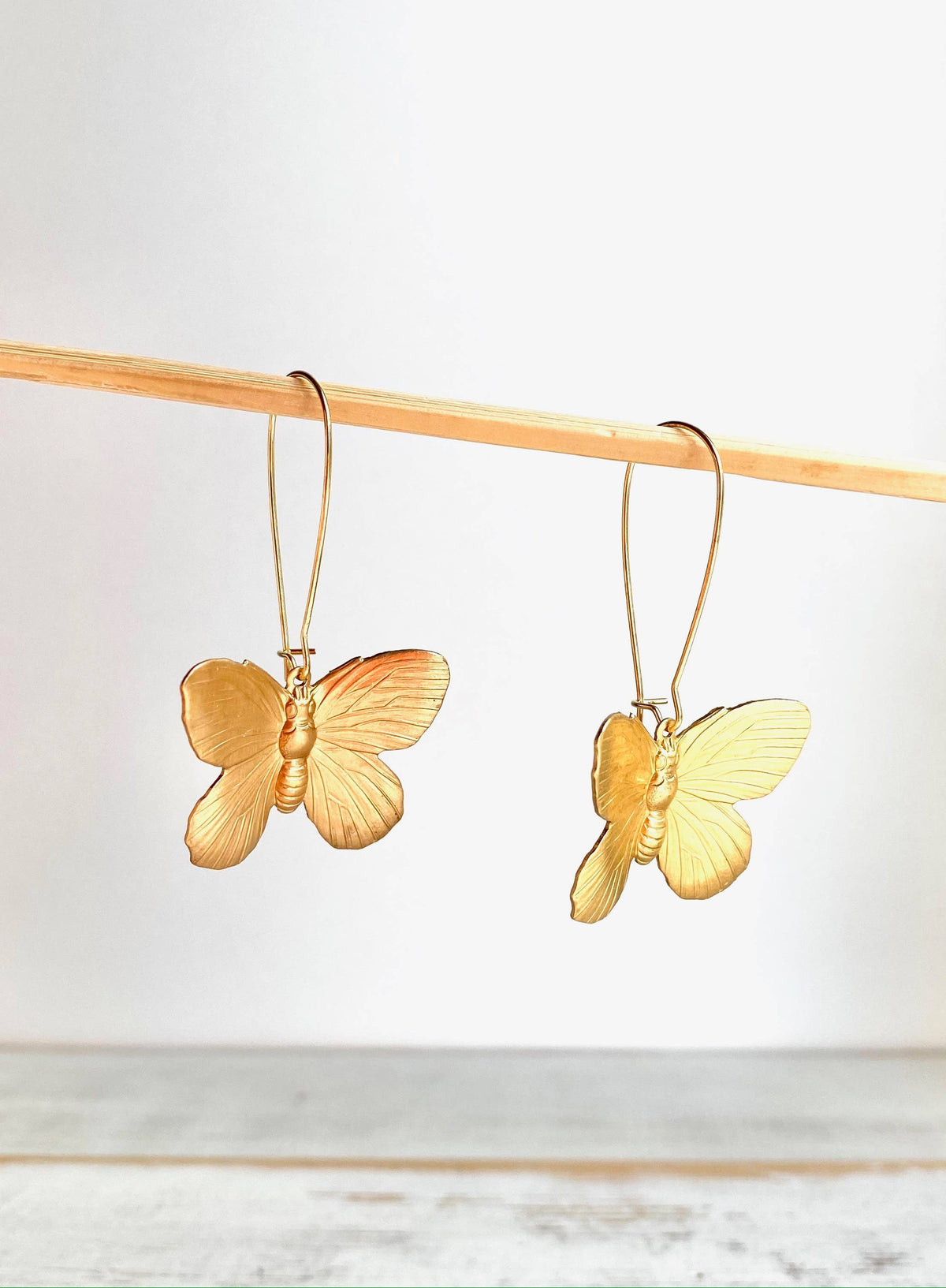 Butterfly Earrings hang from a thin wooden rod against a white background. These golden brass earrings feature delicately detailed butterfly wings with a subtle shine, making them the perfect statement accessory for any occasion.