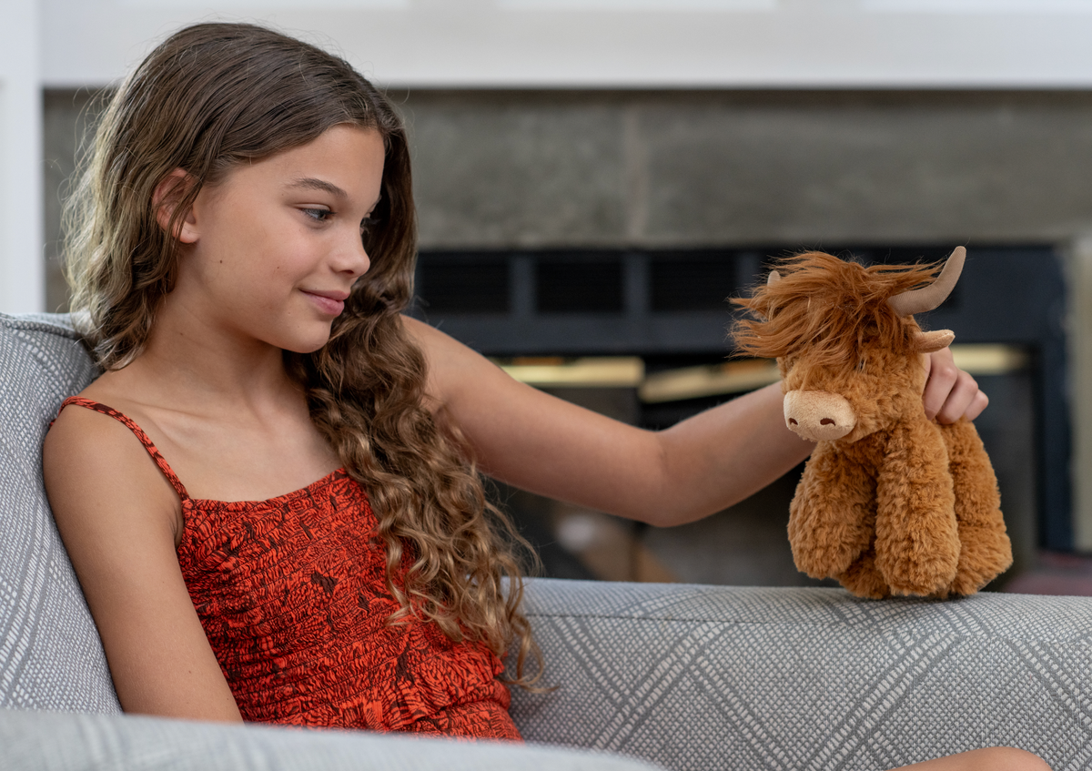 A young girl in a red dress sits on a couch, smiling while holding the Wild Onez Highland Cow Small 9″ (20cm) Farm/Barnyard Animal plush.