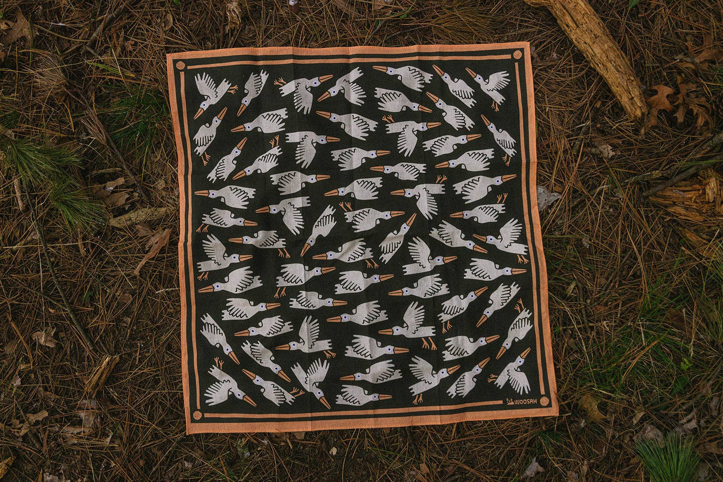 The Migration Bandana is a square cotton accessory with a Woosah Bandanas sublimated print: white birds with orange beaks and feet fly in various directions on a black background, bordered in brown, lying on a forest floor.