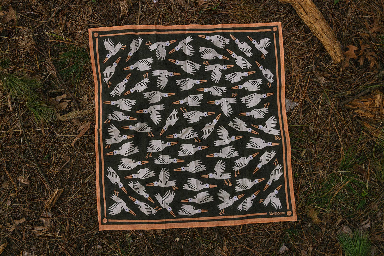 The Migration Bandana is a square cotton accessory with a Woosah Bandanas sublimated print: white birds with orange beaks and feet fly in various directions on a black background, bordered in brown, lying on a forest floor.