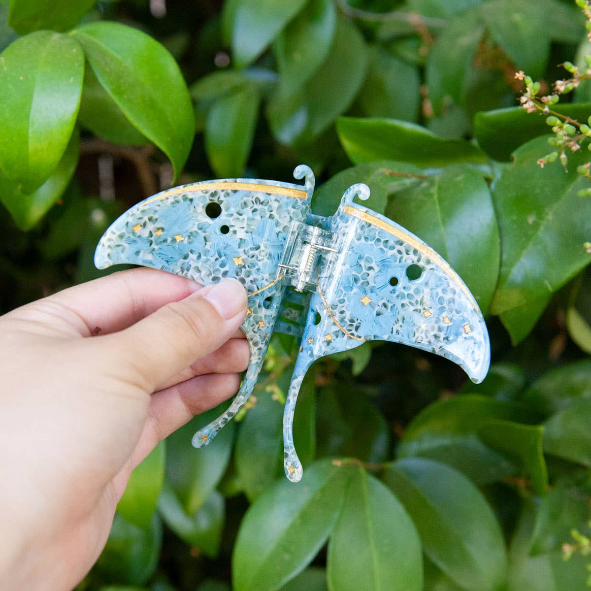 A hand holds a blue acetate Luna Moth Hair Claw Clip in the shape of a luna moth, featuring gold star and line accents.
