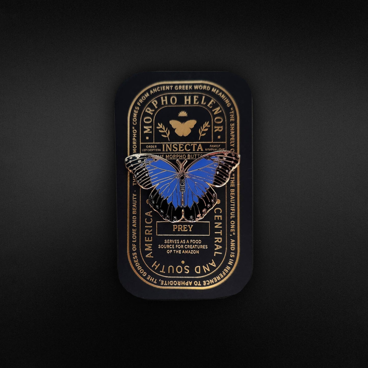 Showcase your appreciation for nature with the Blue Morpho Butterfly Educational Insect Enamel Pin. This elegant black tin display features a striking blue butterfly, gold decorative text, and ornate borders. The highlighted text identifies the Morpho Helenor as a Central and South American species and describes its significance as prey within the food chain. Perfect for collectors and enthusiasts, this beautifully designed pin makes a thoughtful nature gift or unique addition to any butterfly collection.
