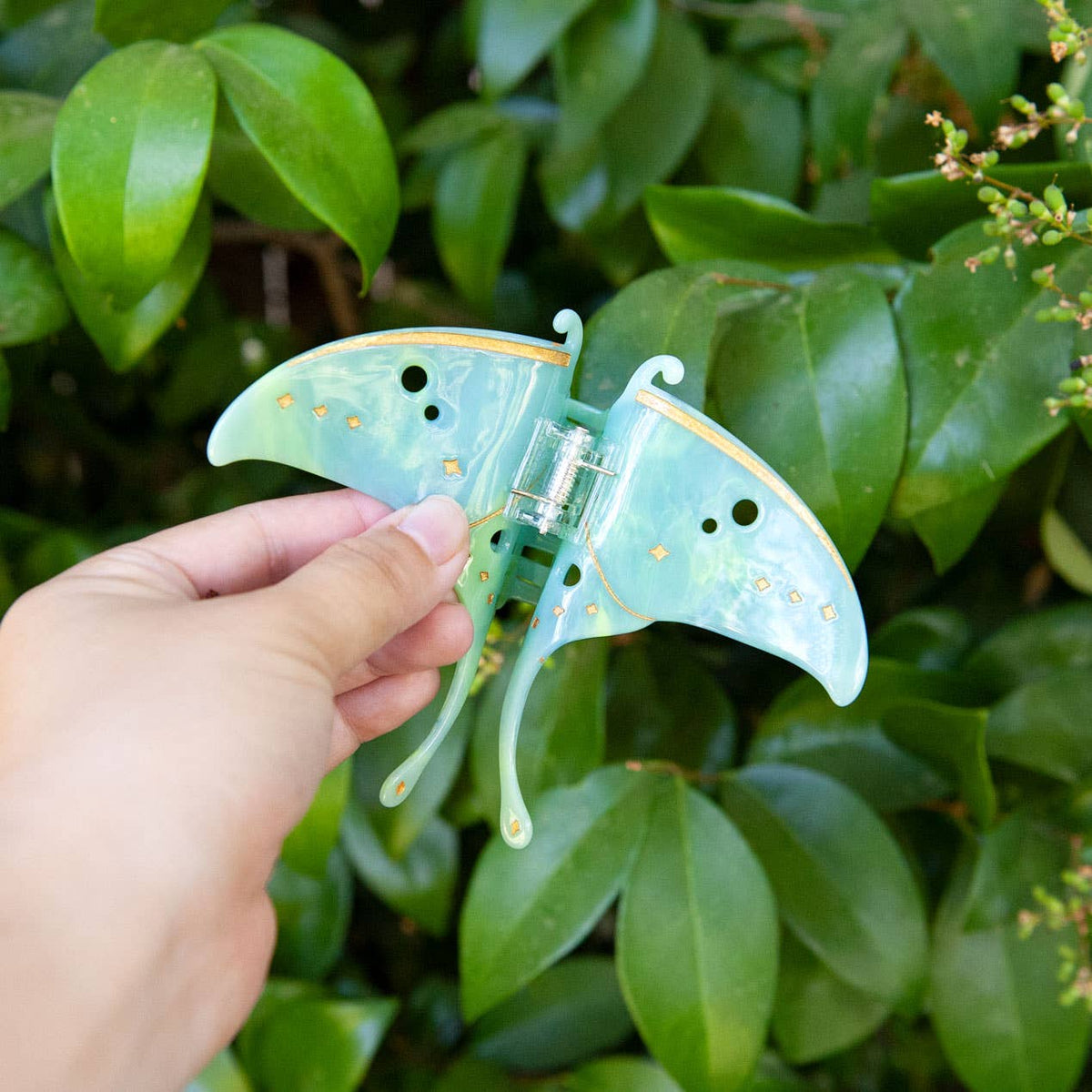 A hand holds a mint green Luna Moth Hair Claw Clip with gold detailing against a background of green leaves.