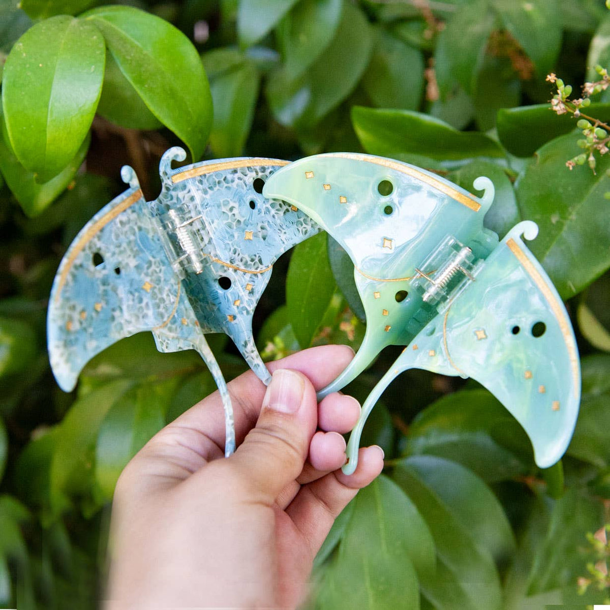 A hand holds two Luna Moth Hair Claw Clips in moth-shaped acetate, one mottled blue and the other translucent light green, both with gold detailing.
