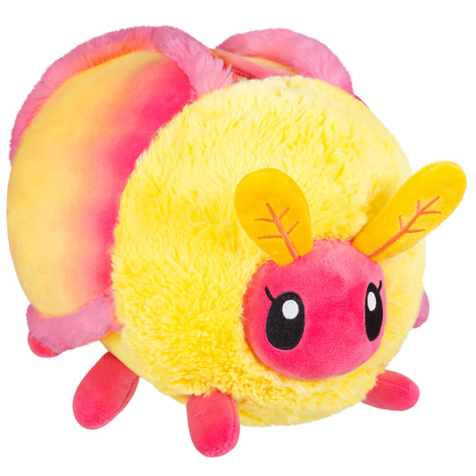 The Mini Squishable Rosy Maple Moth is a plush toy with yellow and pink fur, big black eyes, small antennae, short legs, a soft texture, and a cute round shape inspired by the rosy maple moth.