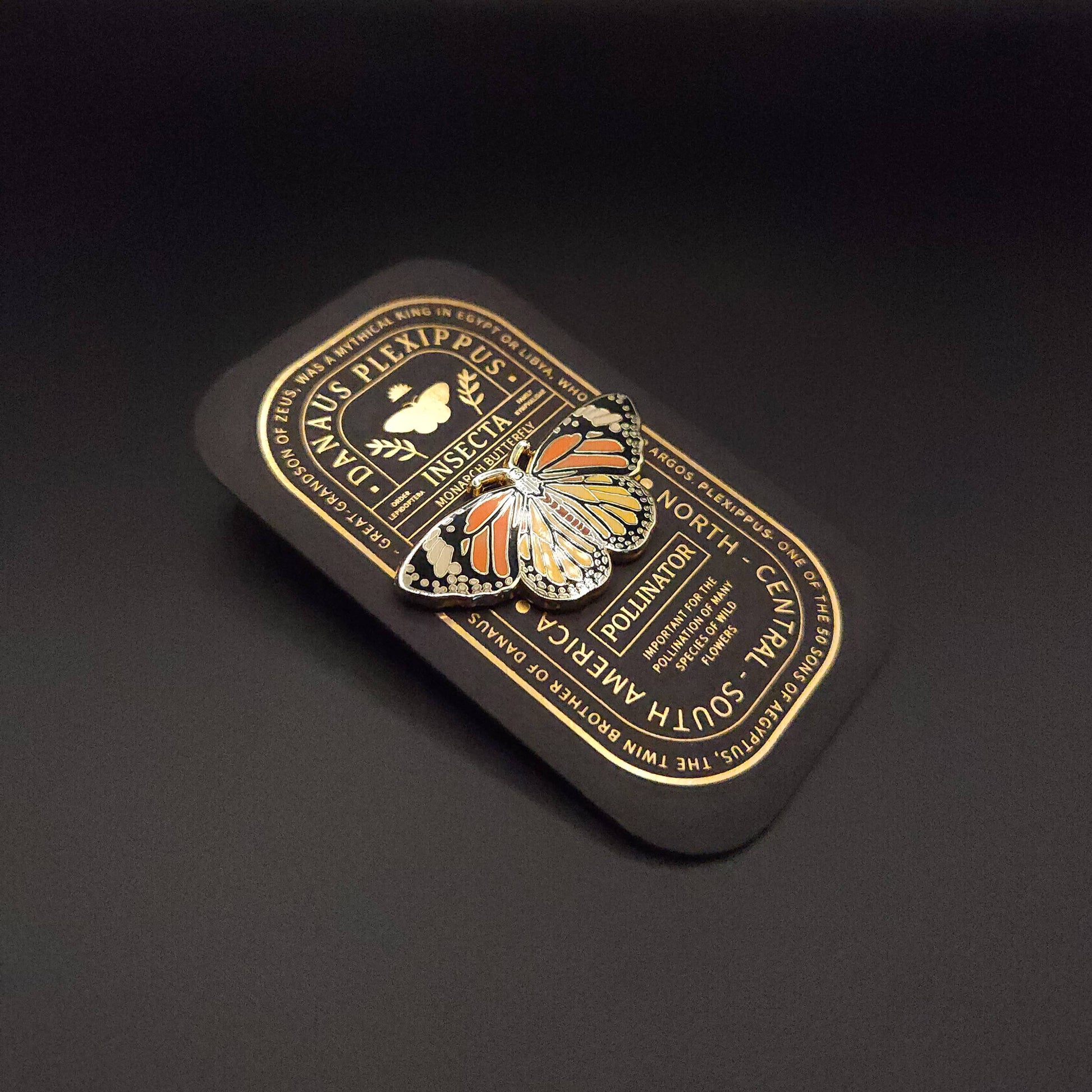 A vibrant Monarch Butterfly Educational Insect Enamel Pin, featuring striking orange, black, and white wings, rests on a rectangular black card adorned with elegant gold text and a small insect illustration at its center. This exquisite hard enamel pin makes a beautiful addition to any butterfly jewelry collection.