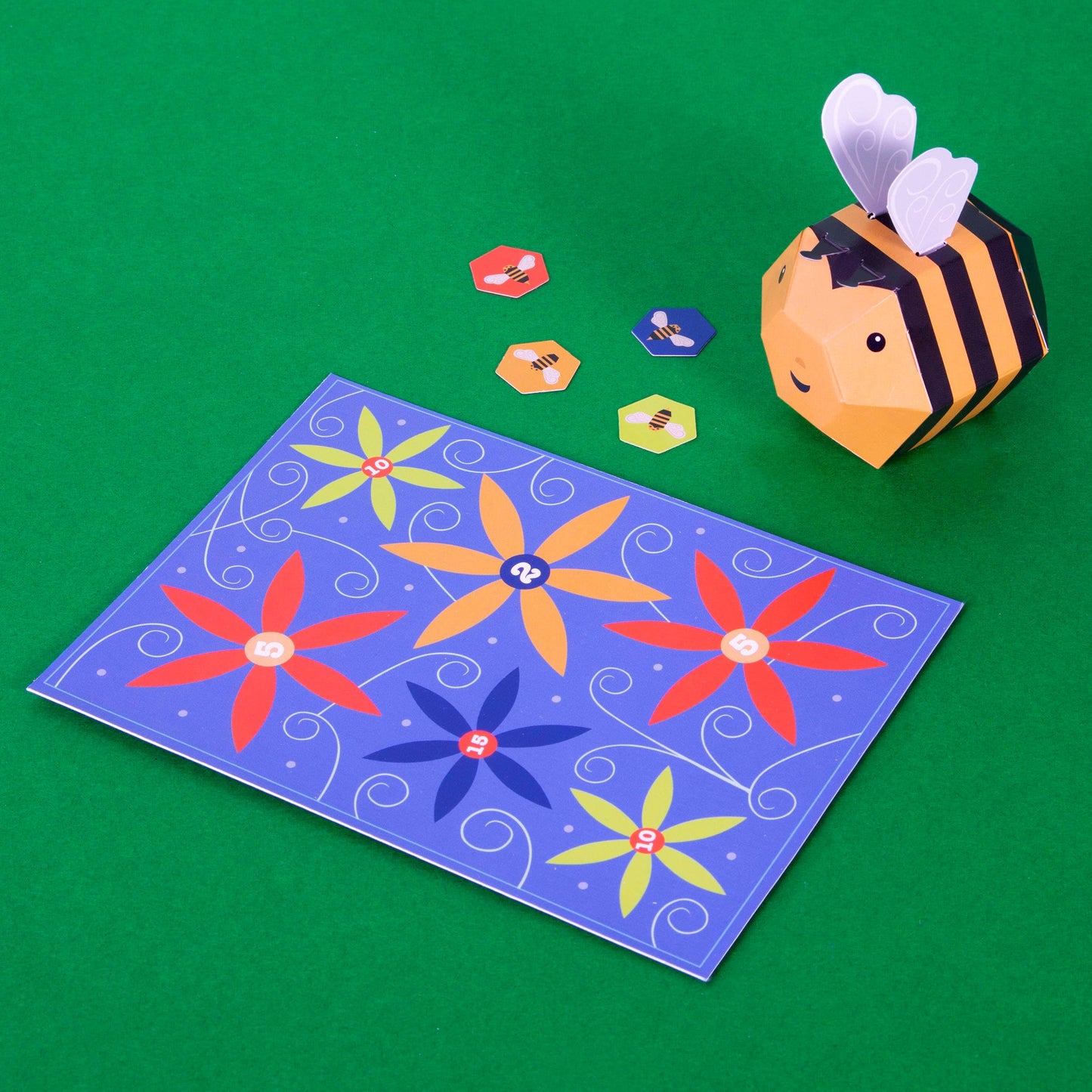 Create Your Own Buzzy Bumble Bee is a plastic-free craft kit with hexagonal tokens featuring bee designs and a blue pollination game board decorated with colorful flowers and numbers, all displayed on a green surface.
