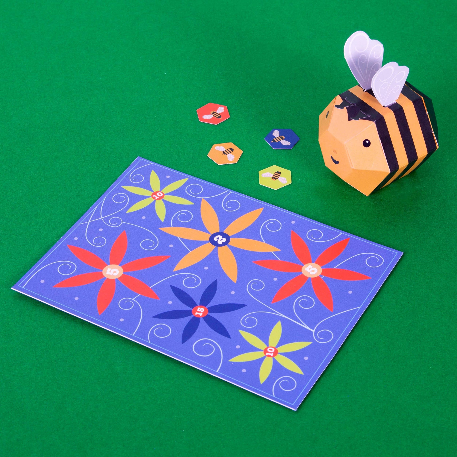 Create Your Own Buzzy Bumble Bee is a plastic-free craft kit with hexagonal tokens featuring bee designs and a blue pollination game board decorated with colorful flowers and numbers, all displayed on a green surface.