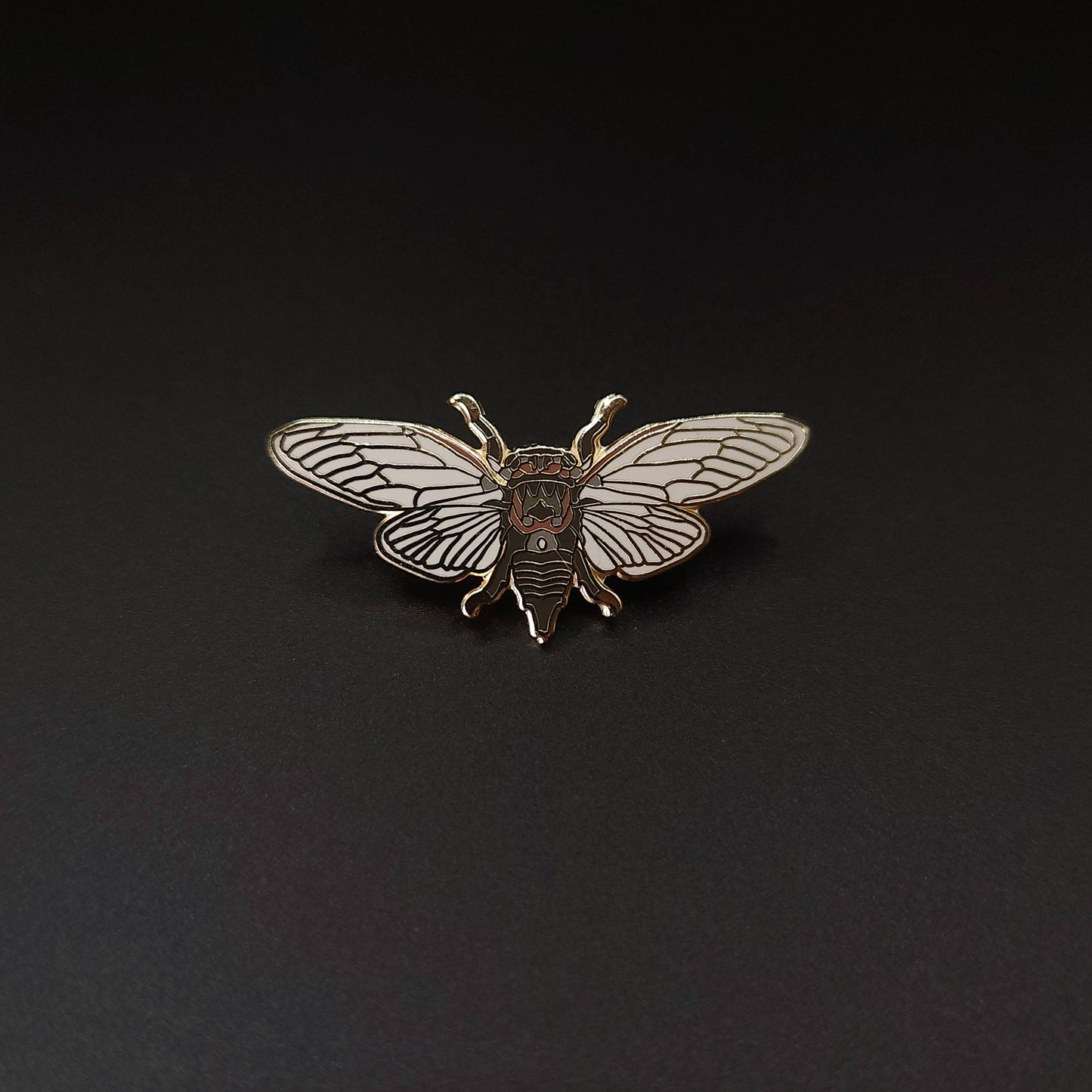 A gold and white hard enamel pin shaped like a cicada rests on a plain black background, making the Scissor Grinder Cicada Educational Insect Enamel Pin a perfect addition to any entomology gift collection.