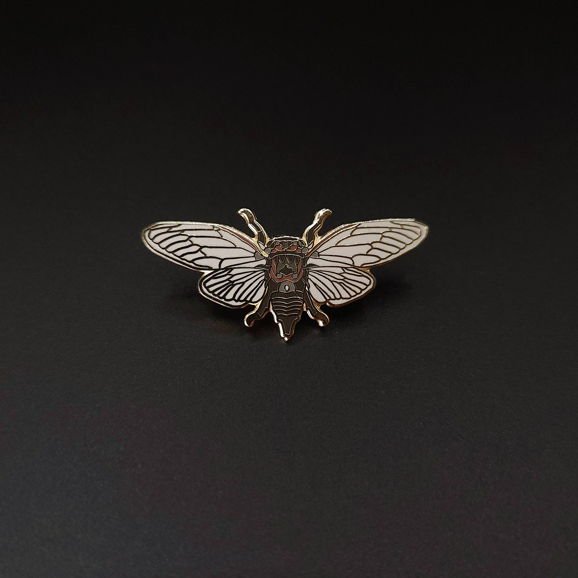 A gold and white hard enamel pin shaped like a cicada rests on a plain black background, making the Scissor Grinder Cicada Educational Insect Enamel Pin a perfect addition to any entomology gift collection.