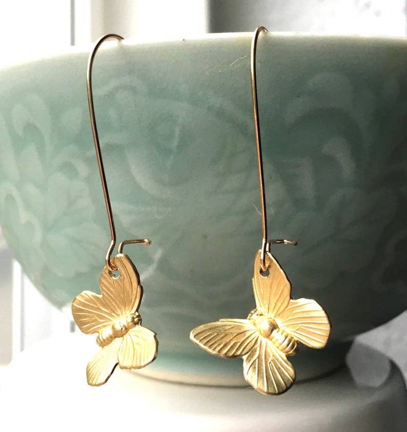 A pair of SmallButterfly Earrings, featuring delicate gold butterfly charms with long hooks, are displayed hanging from the rim of a light blue patterned bowl.