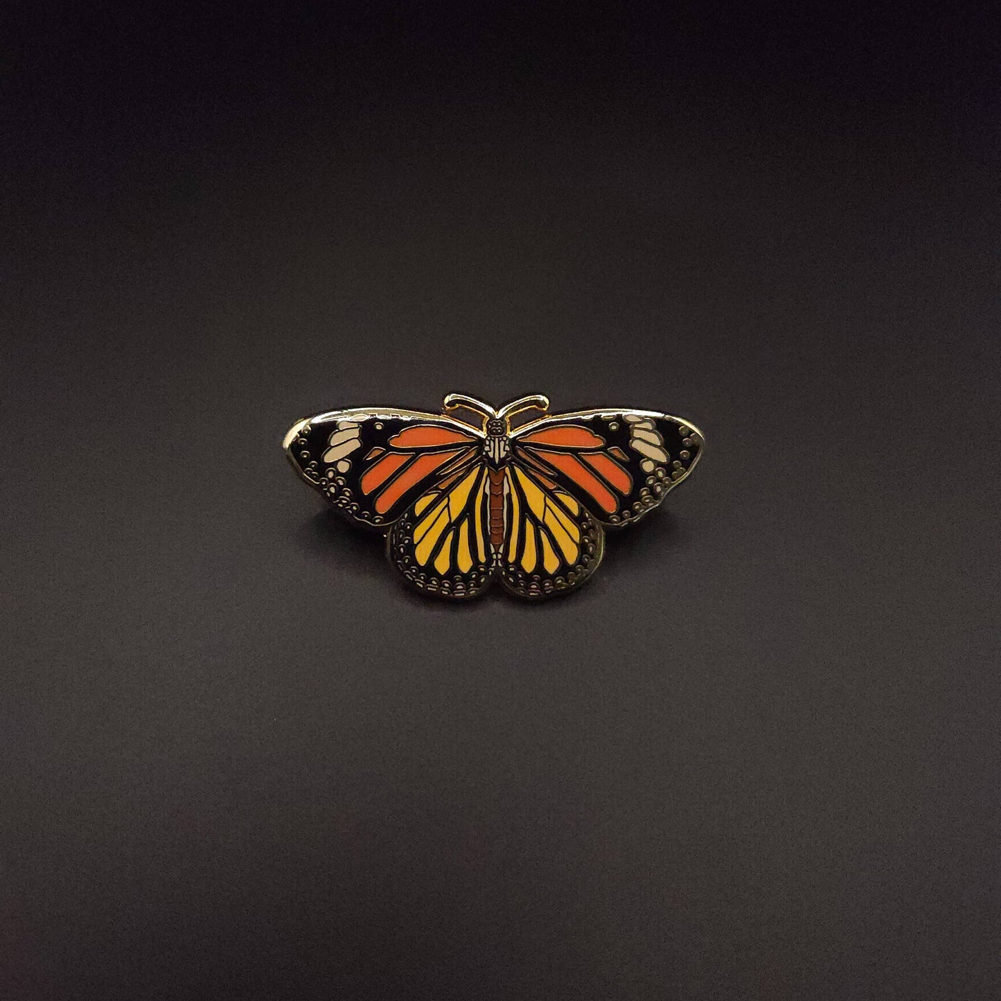 The Monarch Butterfly Educational Insect Enamel Pin, featuring vibrant orange, black, and white details, is centered against a plain black background.