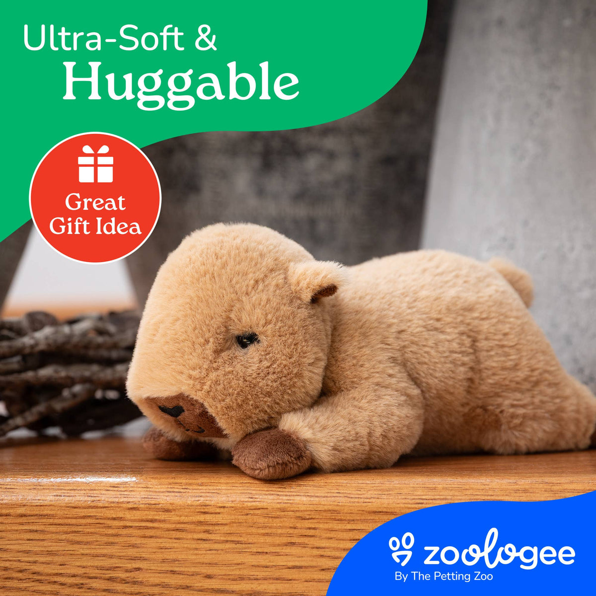 A Scoopz Capybara Small 8″ (21cm) plush toy in light brown lies on a wooden surface, featuring “Ultra-Soft & Huggable” and “Great Gift Idea” text along with the Zoologee logo in the corner—perfect as a stuffed animal capybara for safari nursery decor.