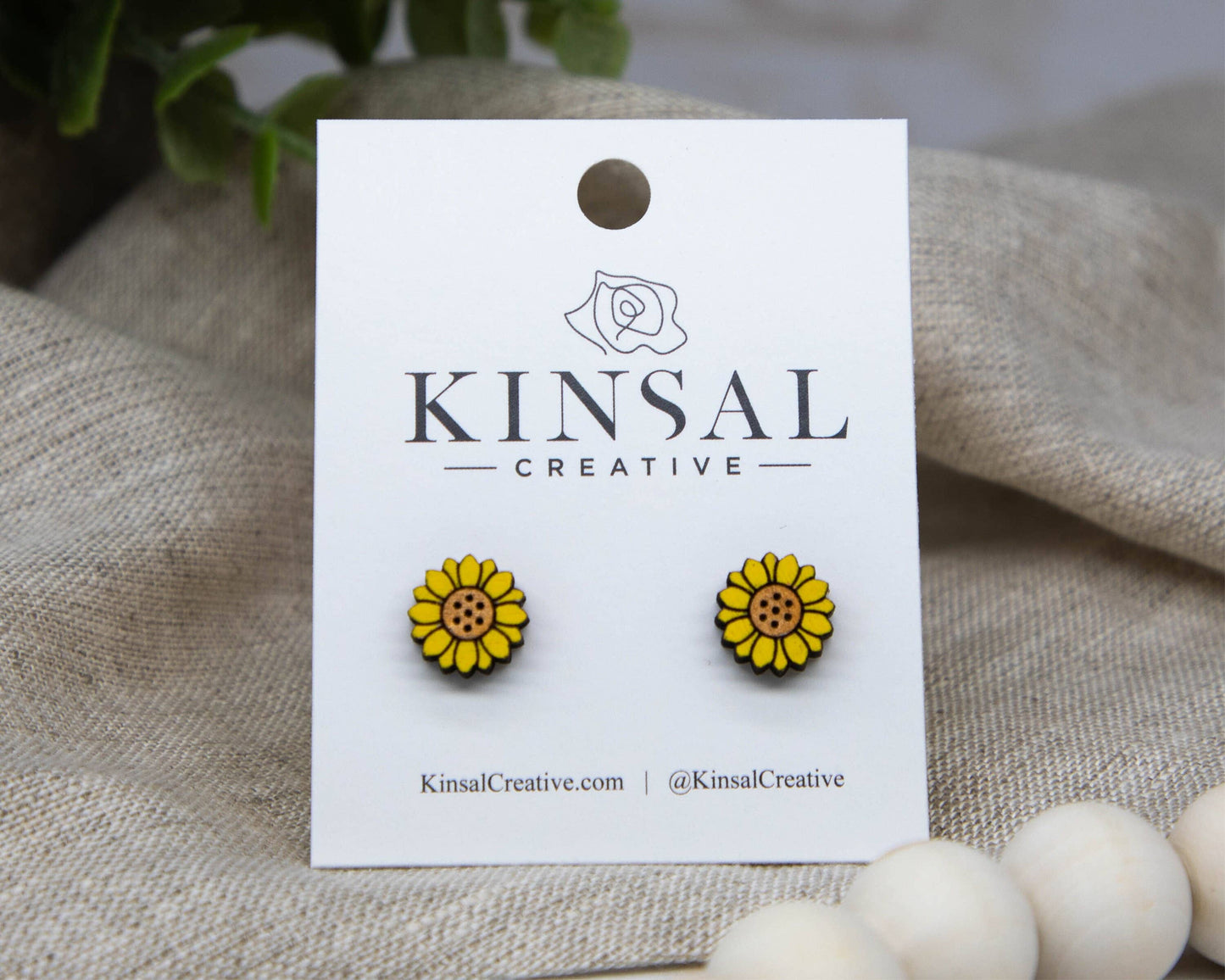 A pair of Sunflower Wood Stud Earrings displayed on a white card labeled Kinsal Creative, surrounded by neutral fabric and wooden beads.