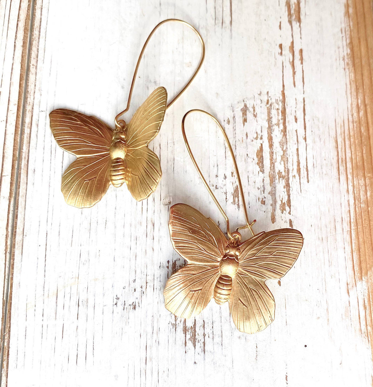 Two Butterfly Earrings in golden brass, featuring detailed wings and long hooks, are displayed on a rustic white wooden surface, making them the perfect statement accessory for any occasion.