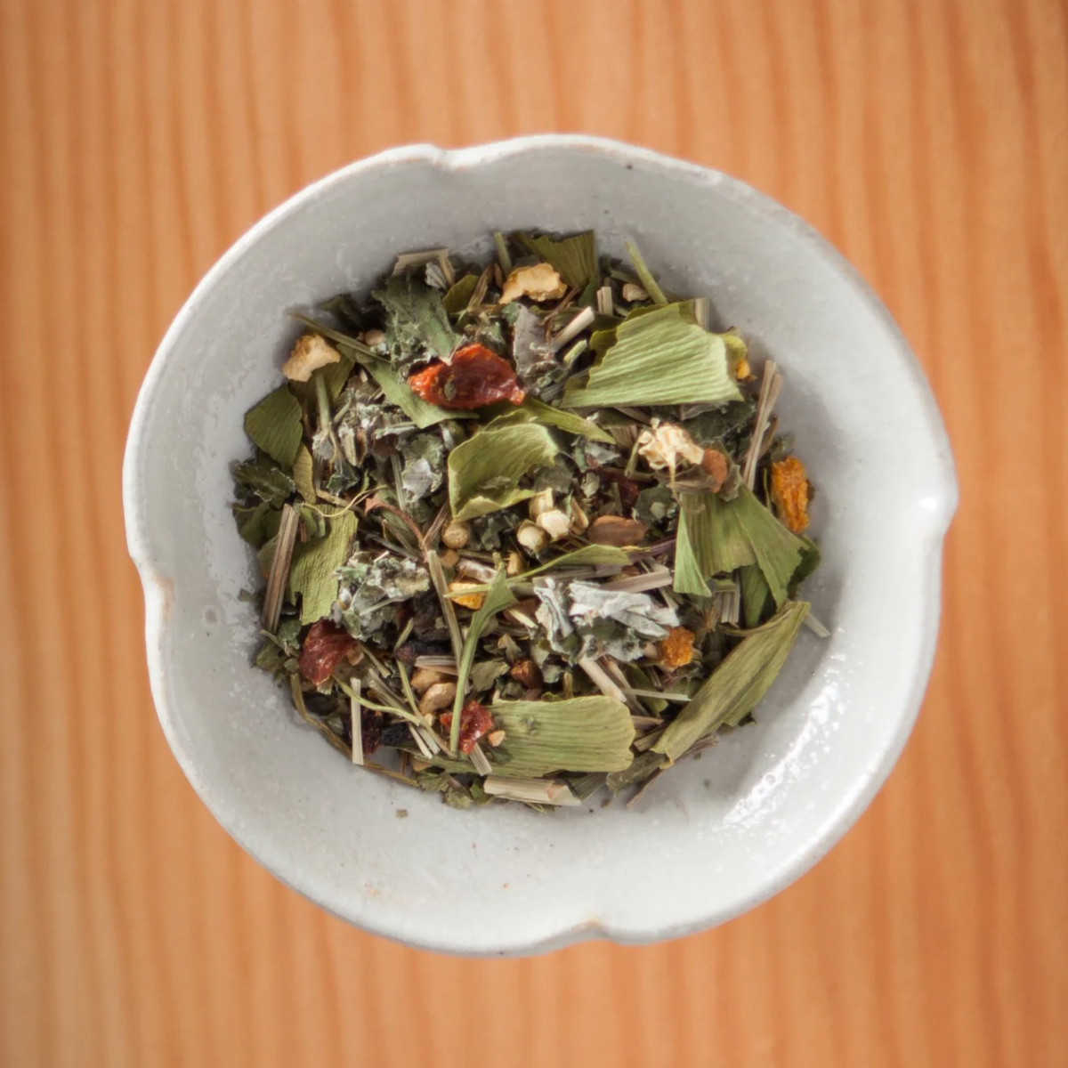A small, white ceramic bowl filled with an organic Bluebird Morning (MWOI) – Loose Leaf Tin tea blend of dried loose-leaf herbs and red fruit pieces.