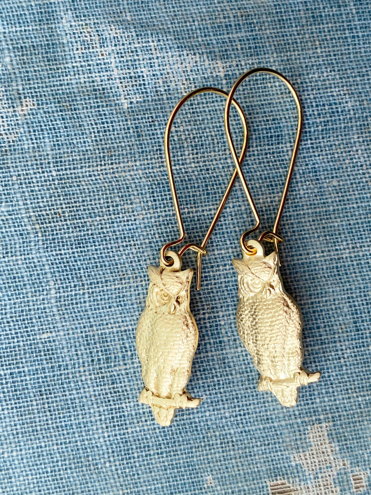 A pair of Wise Owl Earrings featuring vintage brass owls with long hooks, displayed on blue textured fabric adorned with white patterns.