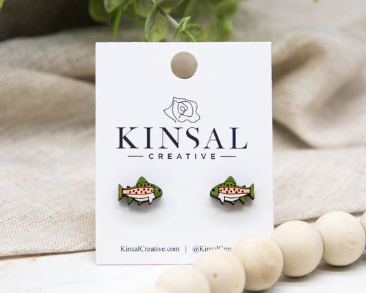 Trout Fish Wood Stud Earrings with green and pink details are showcased on a white "Kinsal Creative" card amid fabric, leaves, and wooden beads—unique handmade jewelry suited for any occasion.