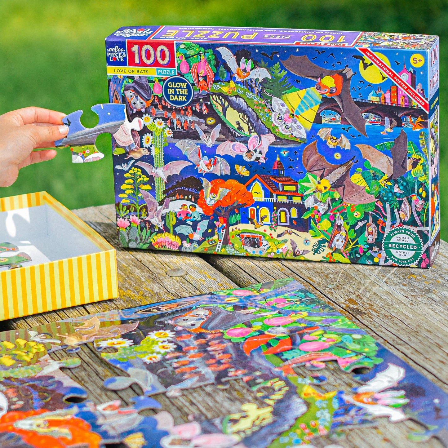 A hand holds a puzzle piece near the completed Love of Bats 100 Piece Glow in the Dark Puzzle and its box, which features vibrant illustrations of bats, plants, and animals. All are arranged on a wooden table.