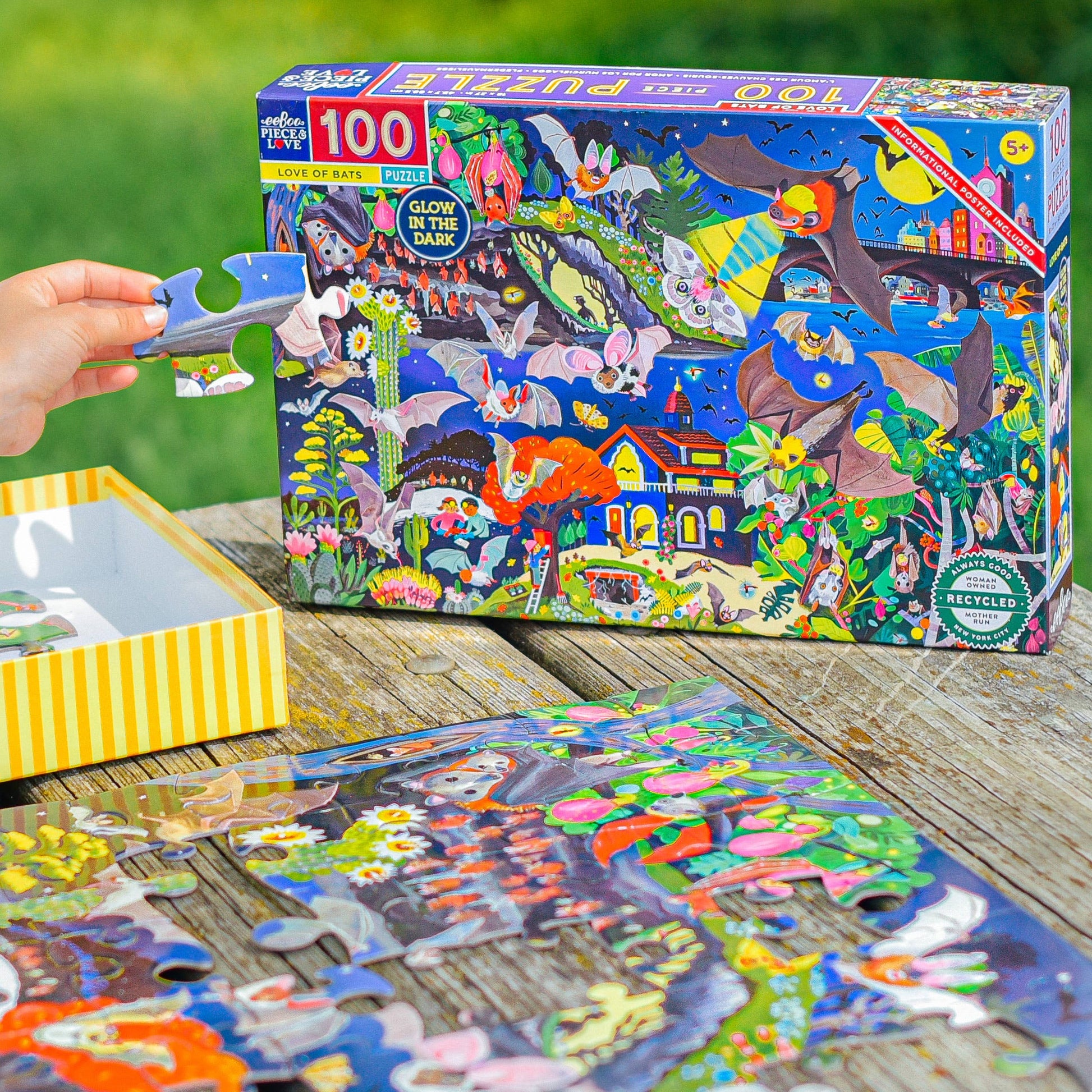 A hand holds a puzzle piece near the completed Love of Bats 100 Piece Glow in the Dark Puzzle and its box, which features vibrant illustrations of bats, plants, and animals. All are arranged on a wooden table.