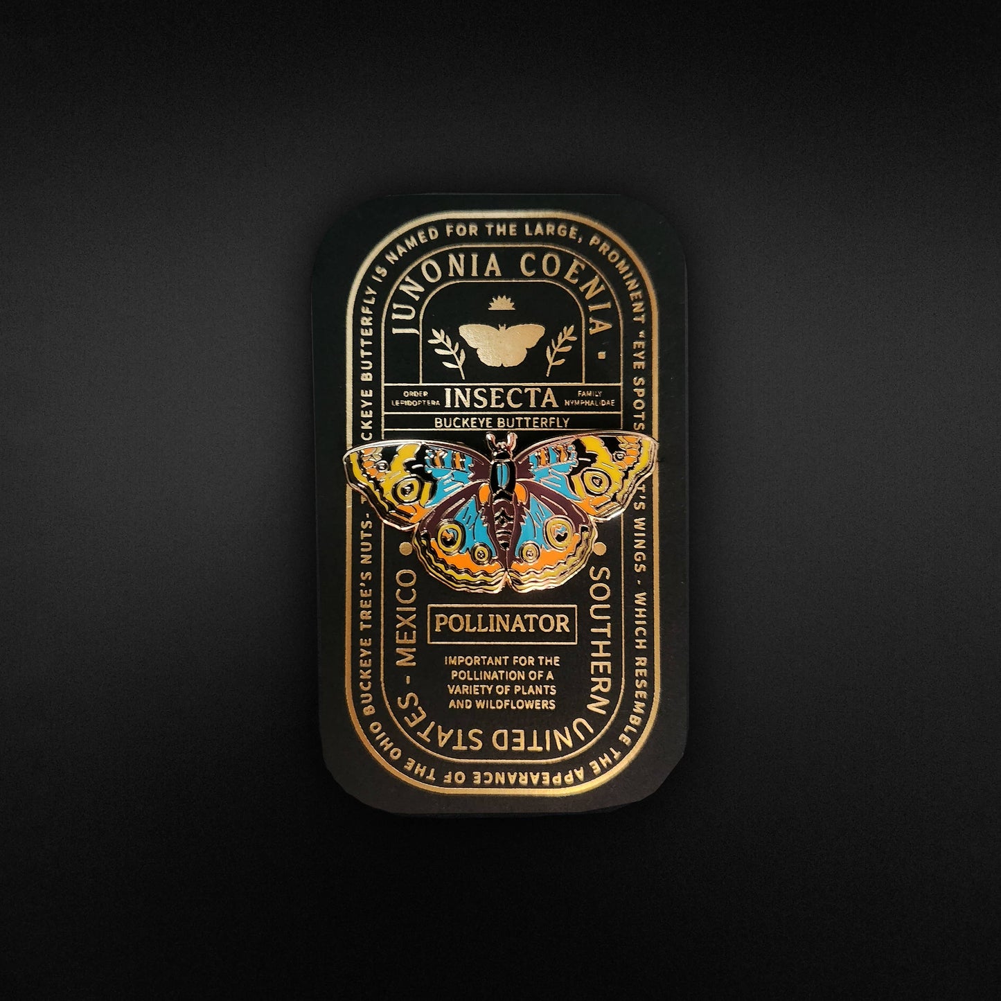 A decorative tin showcases a vibrant butterfly illustration labeled Junonia Coenia, complete with botanical accents and informative text about its importance as a pollinator—making it an ideal gift for butterfly enthusiasts. Ornate gold details set against a black background echo the elegance of the Buckeye Butterfly Educational Insect Enamel Pin, creating an educational and charming display.