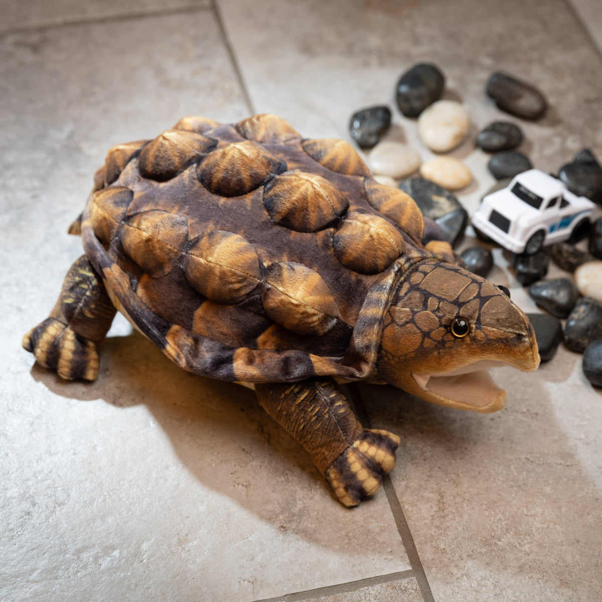 A Wild Onez Snapping Turtle 15″ (38cm) plush sits on a tiled floor near a cluster of rocks and a small white toy car, making it a charming addition to any swamp-themed nursery decor.