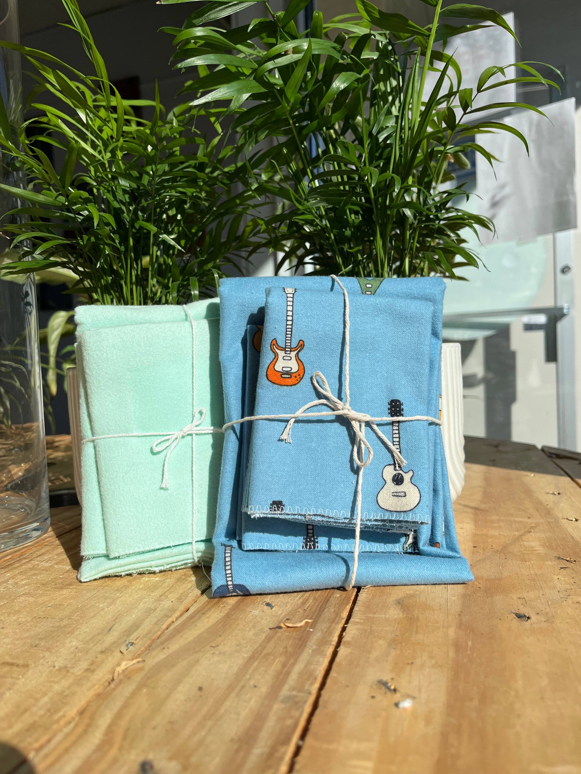 Two small gift-wrapped packages—one pastel green, one blue with guitar prints—tied with white string, sit on a wooden table before potted plants. Perfect for gifting the Dishtowel & 3 Dishcloth Set- Compostable.