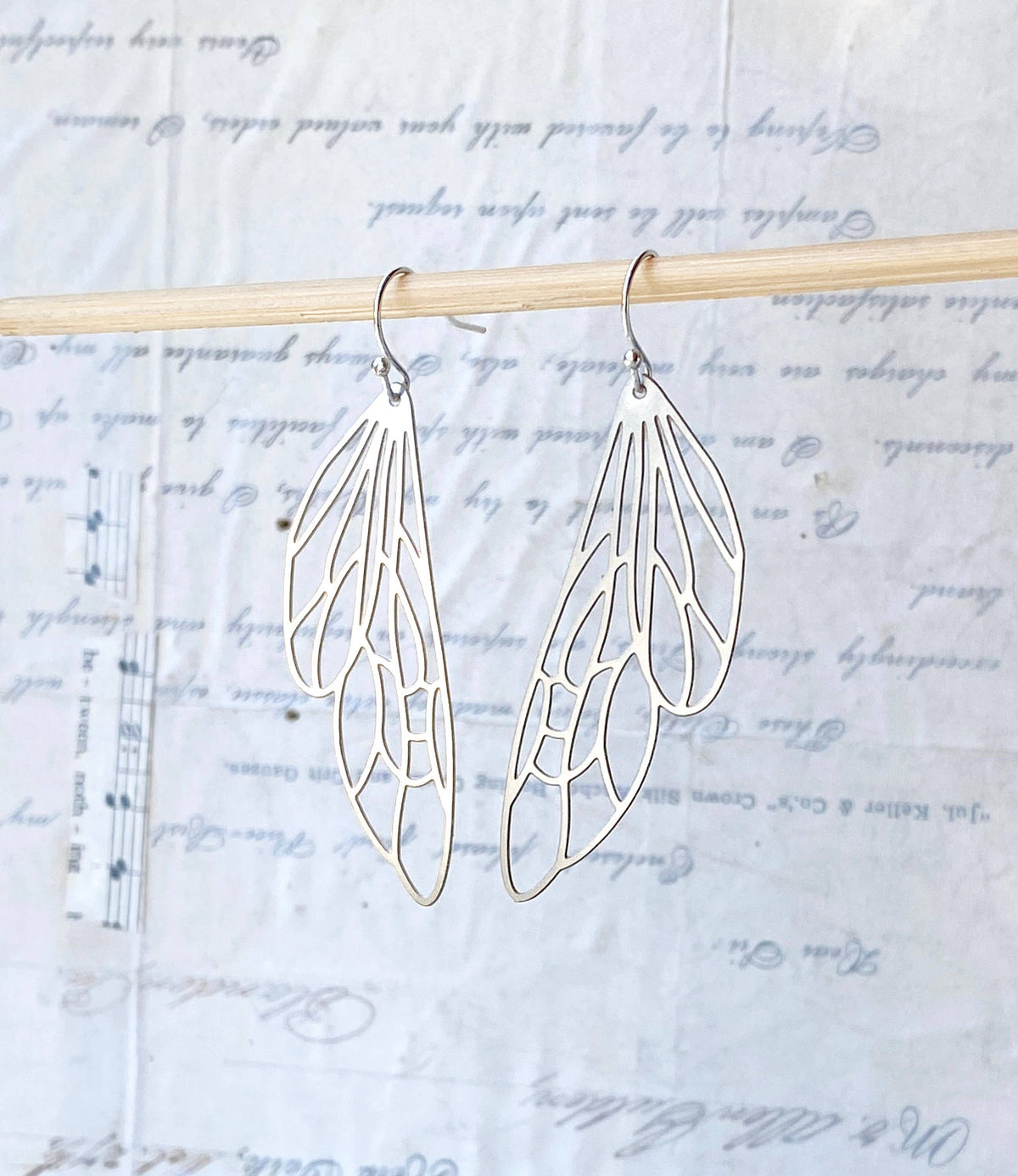 Silver Lace Cicada Wings earrings, shaped like delicate insect wings, hang from a wooden rod against a decorative backdrop featuring handwritten script and faint musical notes.