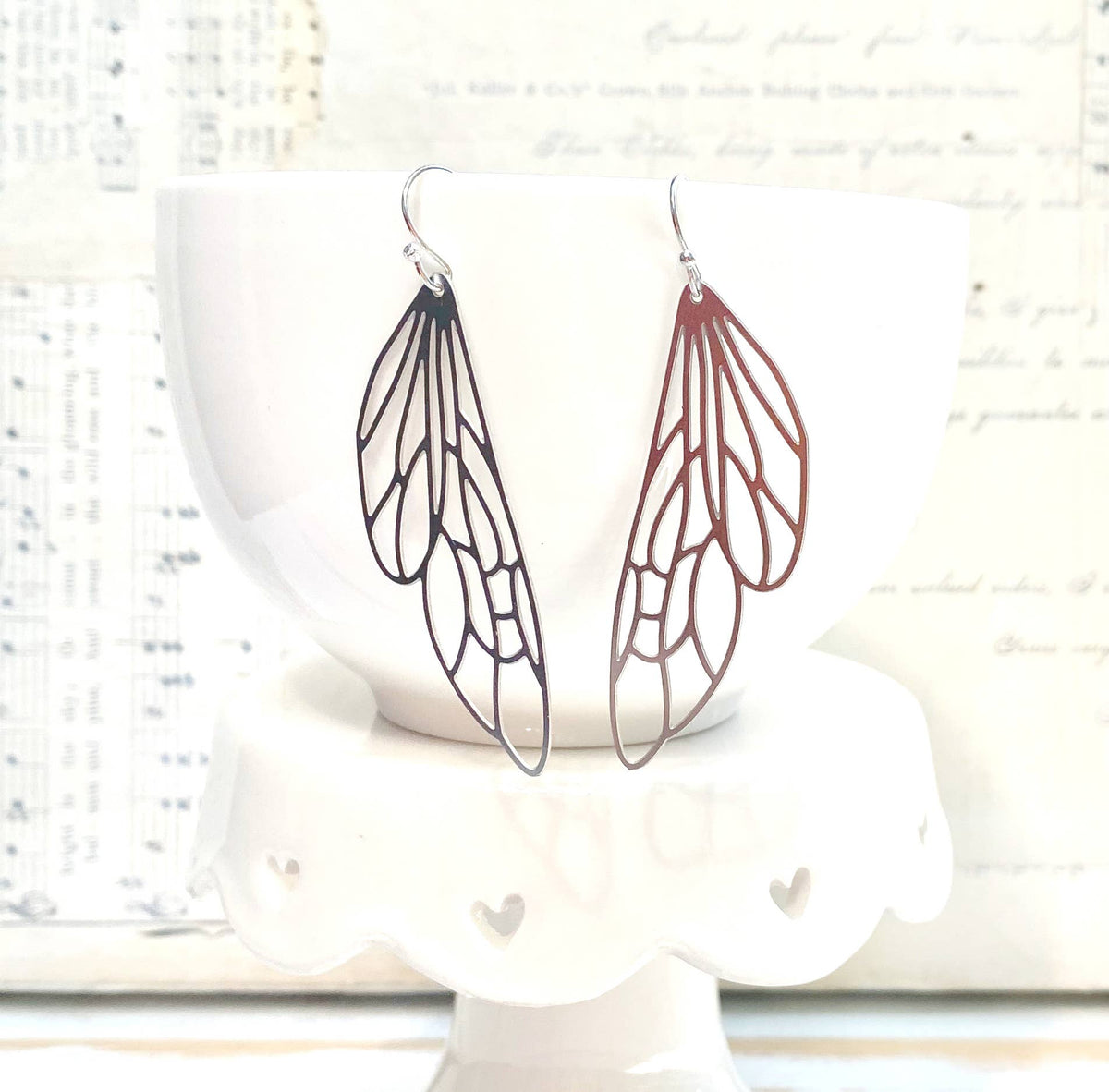 A pair of Silver Lace Cicada Wings earrings, delicately shaped like insect wings, hang from a white bowl resting on a white stand with scalloped edges and small heart cutouts, while blurred sheet music forms the backdrop.