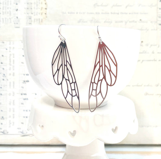 A pair of Silver Lace Cicada Wings earrings, delicately shaped like insect wings, hang from a white bowl resting on a white stand with scalloped edges and small heart cutouts, while blurred sheet music forms the backdrop.