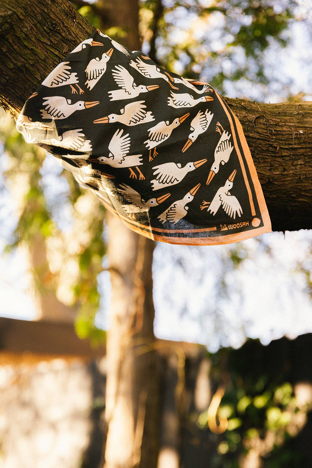 The Migration Bandana, featuring white bird illustrations on patterned cotton, is draped over a tree branch outdoors with sunlight streaming through green leaves—a peaceful scene from Woosah Bandanas.