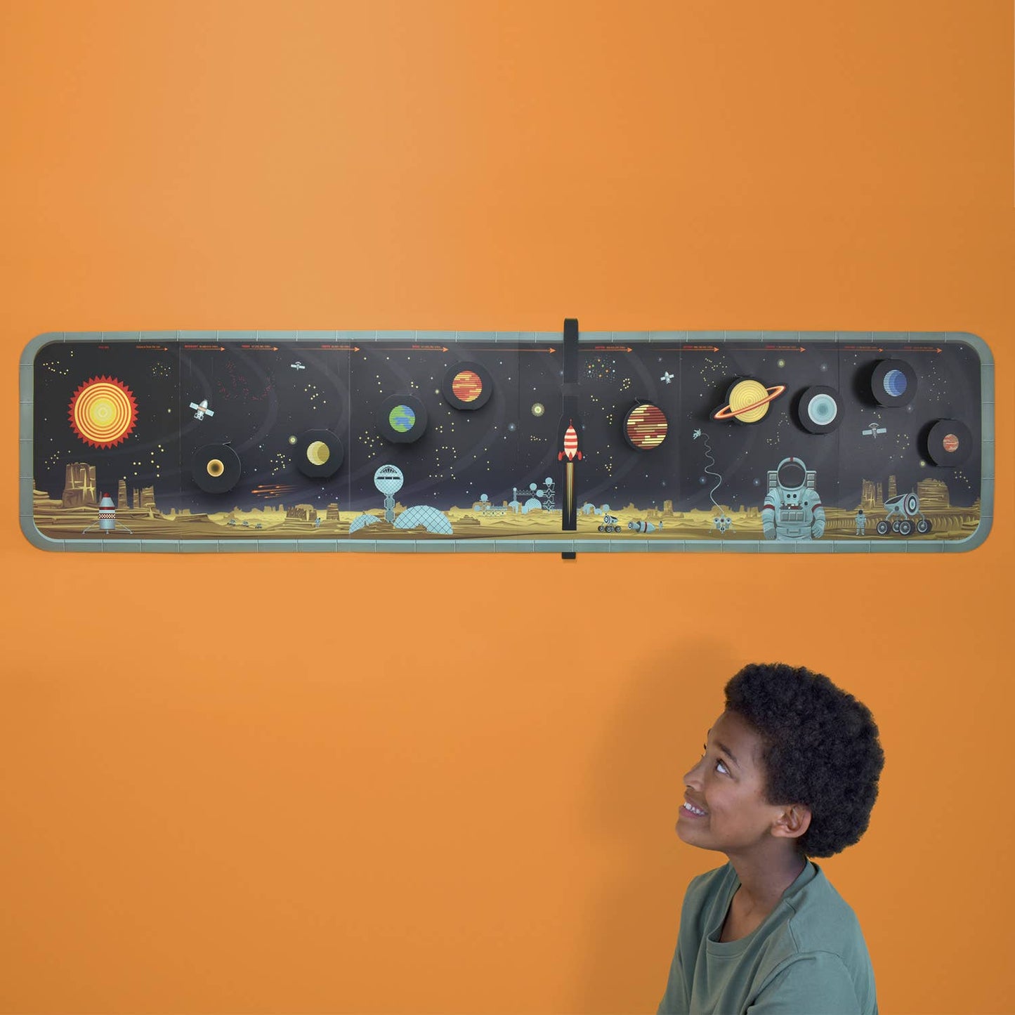 A smiling boy admires Create Your Own Solar System, a 3D wall panel with lifelike planets, rocket, astronaut, and vibrant space scene on an orange wall—an interactive solar system activity kit brought to life.