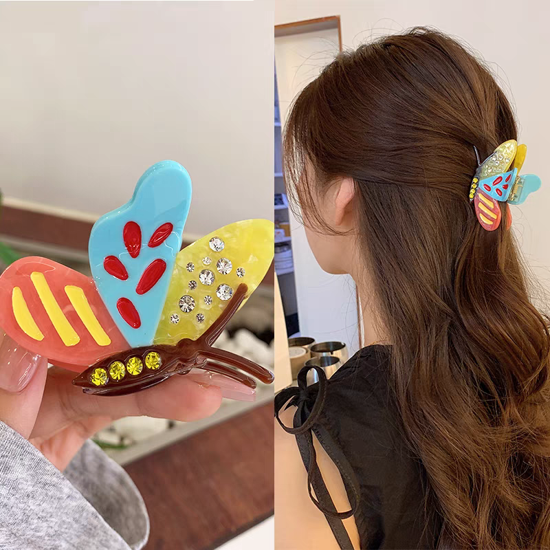 A Side Butterfly Hair Claw Clip with colorful resin wings, rhinestones, and yellow accents, shown held in a hand and worn in brown hair as a stylish accessory.