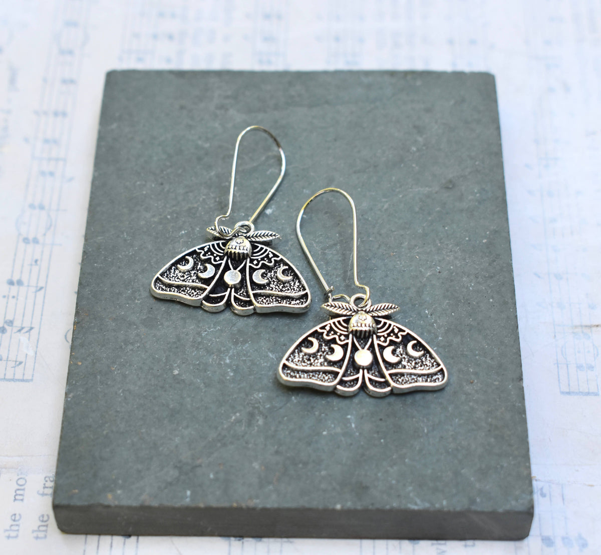 The Luna Moth Earrings, featuring intricate silver detailing, are displayed on a gray stone slab with sheet music in the background.