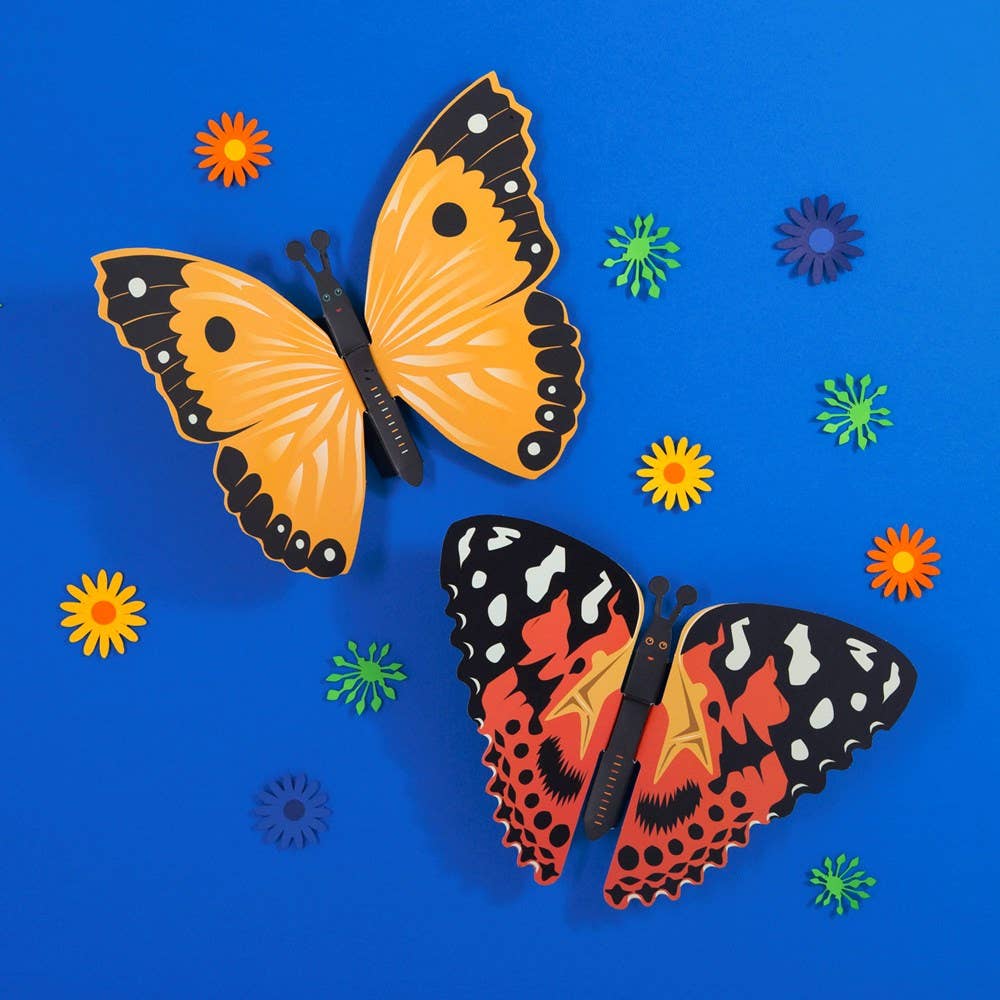 Two colorful paper butterflies from the Create Your Own Fluttering Butterflies kit sit on a blue background with yellow, orange, green, and purple flowers. This no-glue craft is perfect for creative fun.