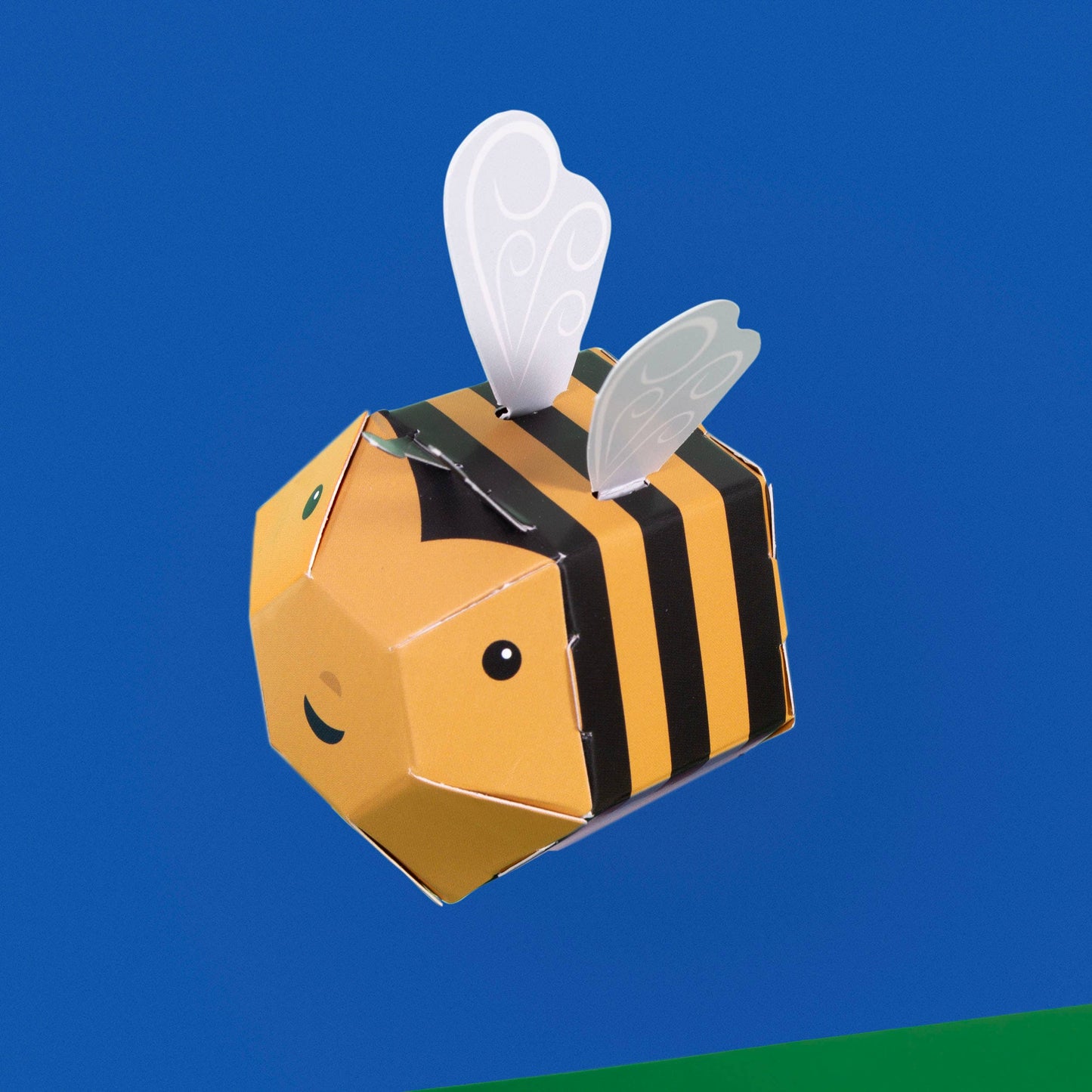 Create Your Own Buzzy Bumble Bee is a plastic-free paper craft featuring a yellow and black striped body, white wings, and a smiling face, set against a blue background with a small green corner at the bottom.