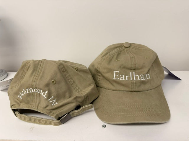 Two khaki Earlham Unstructured Caps featuring white embroidery. One cap displays "Earlham" on the front, while the other features "Richmond, IN" on the back. Both caps are positioned on a white surface against a simple background.