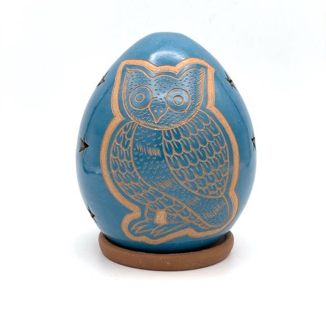 The Owl Luminary is a handcrafted, blue, egg-shaped ceramic ornament with carved owl details and star-shaped cutouts, set on a small round brown base.