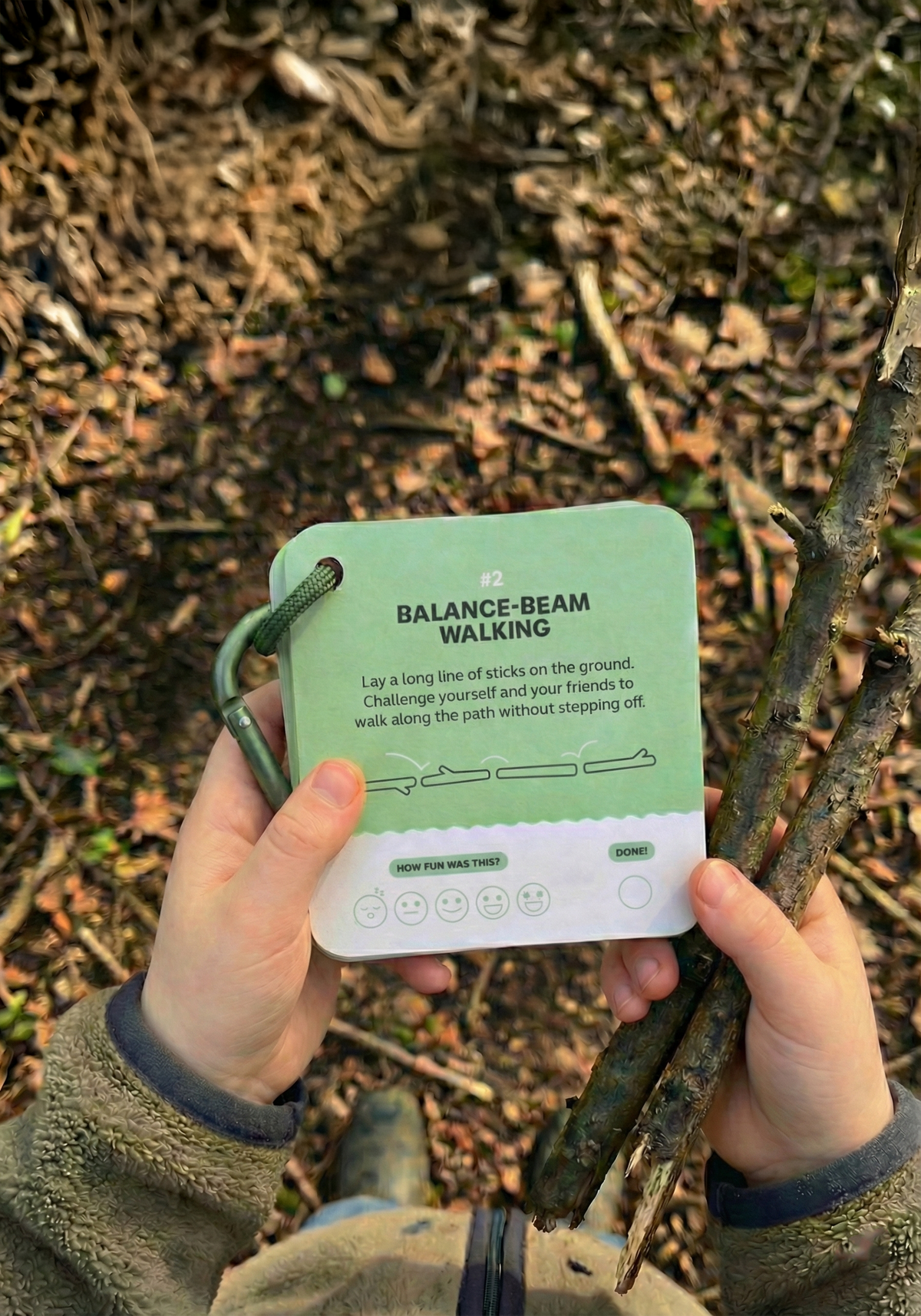 A person holds a stack of Fun Things to do with a stick | Play and Activity Cards, with the top card titled Balance-beam walking showing instructions for a stick-based nature activity.