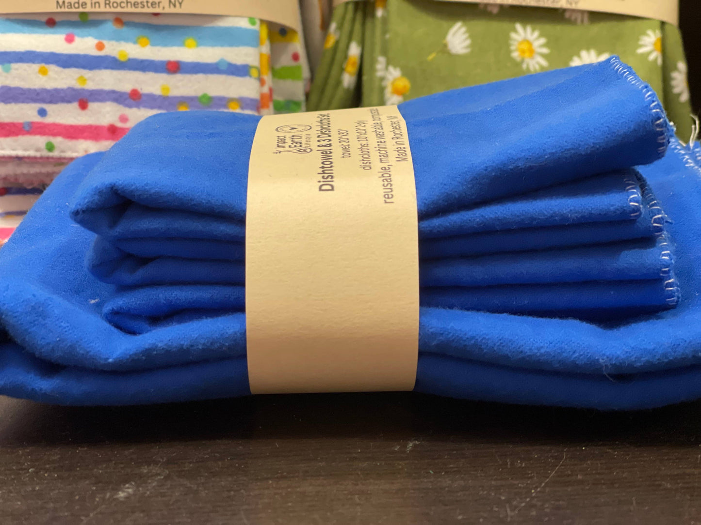 A stack of folded blue cotton flannel from the Dishtowel & 3 Dishcloth Set- Compostable, wrapped with a brown paper band, rests on a dark surface with colorful patterned textiles partially visible in the background.