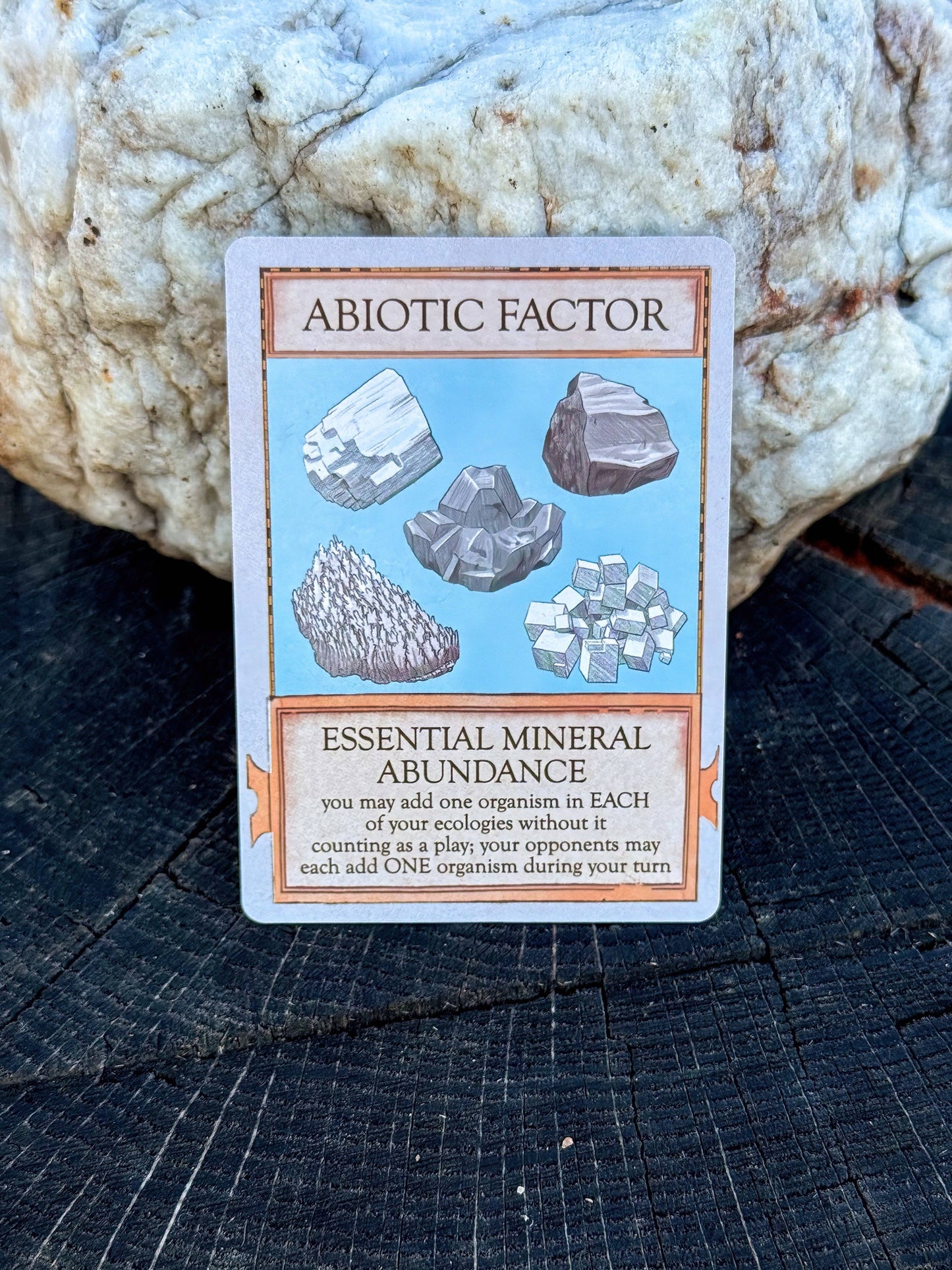 A playing card titled Abiotic Factor, from the scientific card game Ecologies: Marvelous Microbes, features illustrations of six minerals and text describing the Essential Mineral Abundance ability, propped against a white rock on a tree stump.