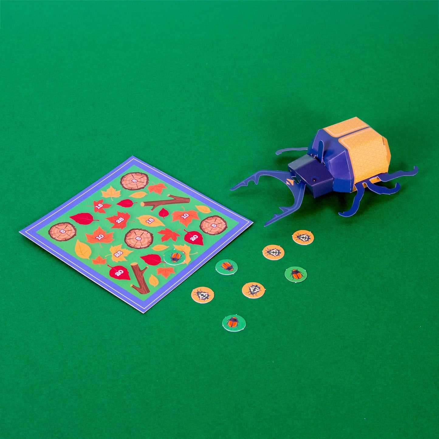 The Create Your Own Super Stag Beetle for Kids, in blue and orange, is shown next to a vibrant game board with leaves, flowers, and round veggie tokens, all arranged on a green surface.