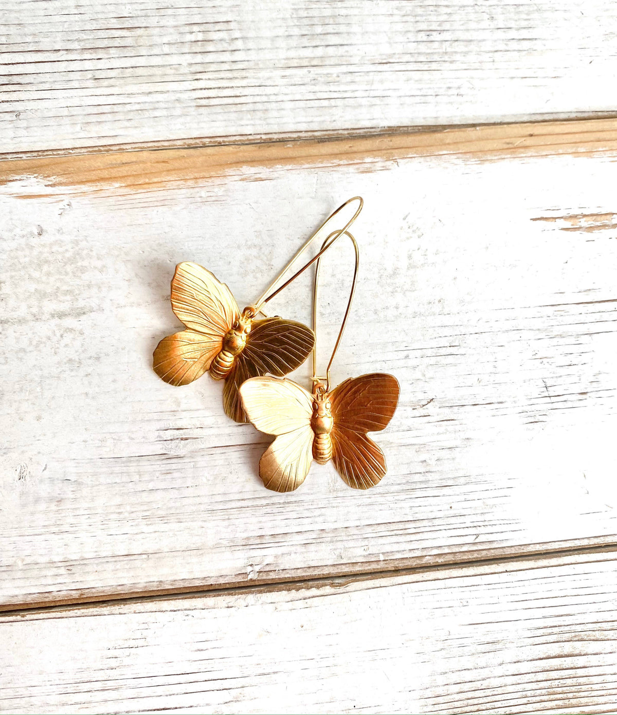 Two Butterfly Earrings featuring fine engraved details are displayed on a rustic white wooden surface.