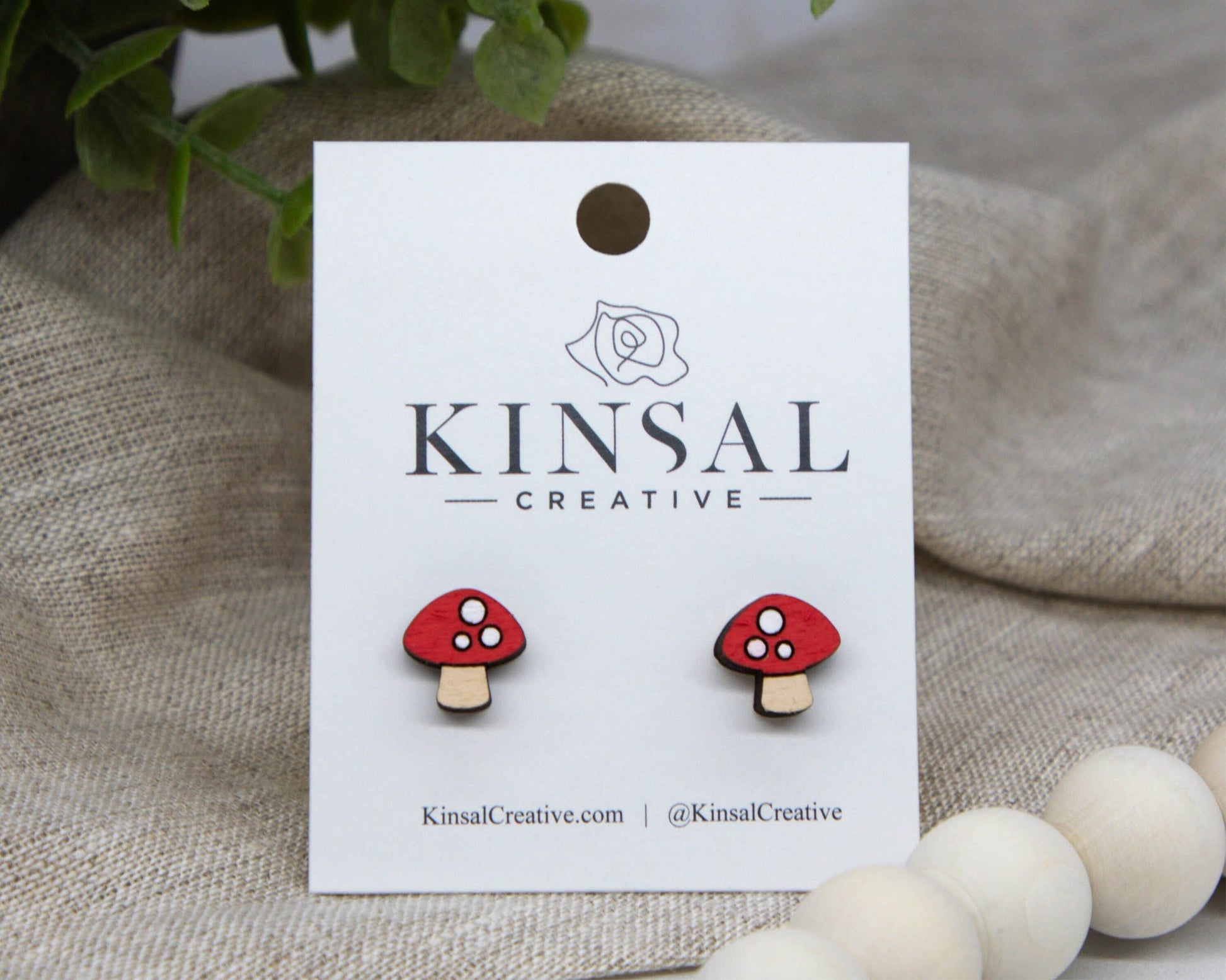 The Mushroom Wood Stud Earrings are handmade, painted red and white to look like mushrooms, displayed on a Kinsal Creative card with a plant and beige fabric in the background—unique jewelry for any collection.